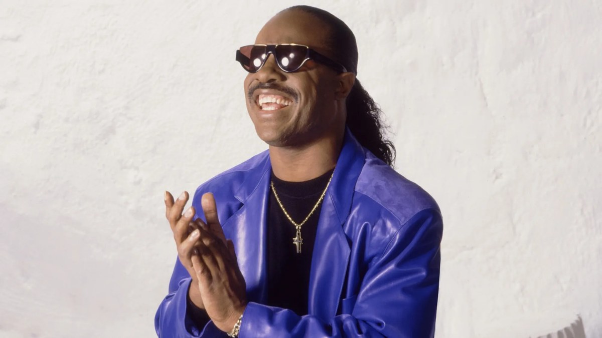 Stevie Wonder smiling in sunglasses and a purple leather jacket, clapping his hands